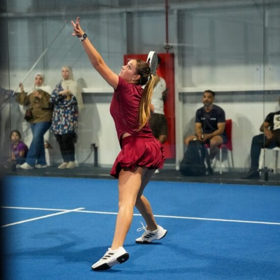 An indoor padel court in play, blue surface under hall lighting.