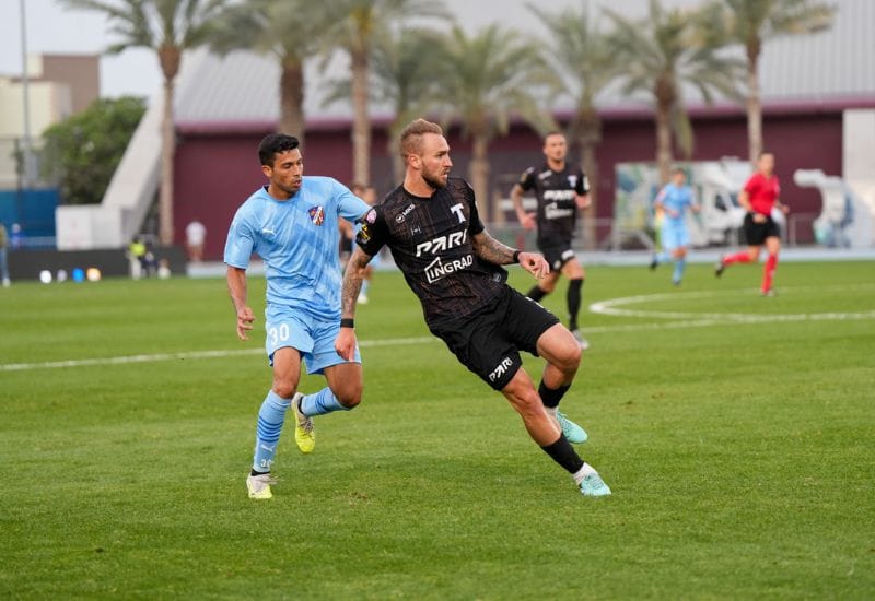 A match on grass with palms behind the pitch.
